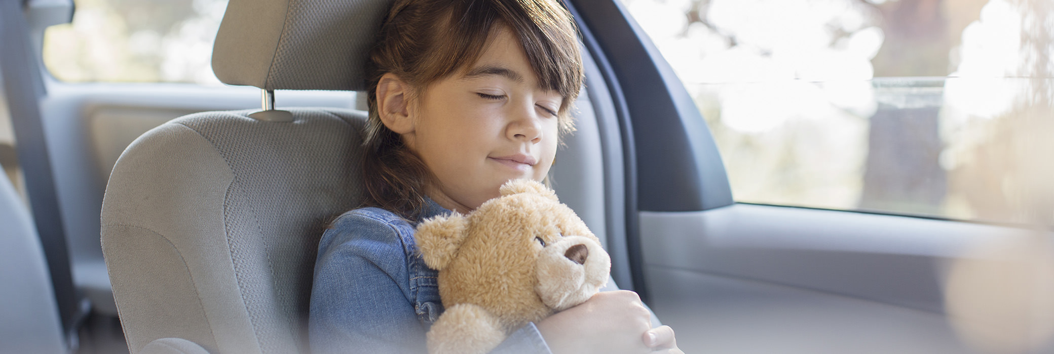 A little girl smiling and sitting in a car hugging her teddy bear with her eyes closed
